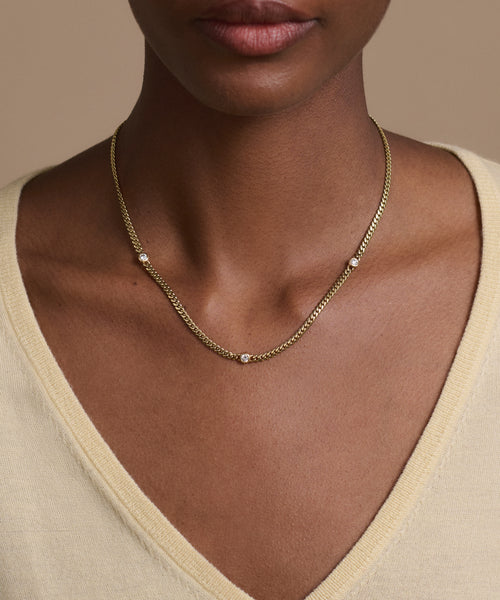 A person wears a light yellow V-neck top and the Merit Round Cut Diamond Necklace, featuring small round diamonds spaced evenly on a 14k gold chain. A neutral beige background highlights the subtle elegance of Jenni Kayne jewelry.