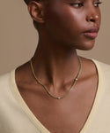 A woman in a light yellow V-neck top wears the Merit Round Cut Diamond Necklace, featuring a delicate diamond pendant, while looking slightly to the side against a beige background.