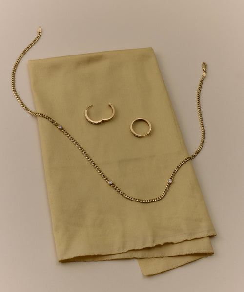 The Merit Round Cut Diamond Necklace from Jenni Kayne jewelry is displayed on folded, light yellow fabric against a beige background.