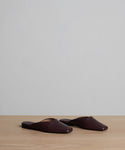 The Mia Mule: a pair of dark brown Italian leather slip-on mules with a pointed toe, displayed side by side on a light wooden surface against a plain light gray background.