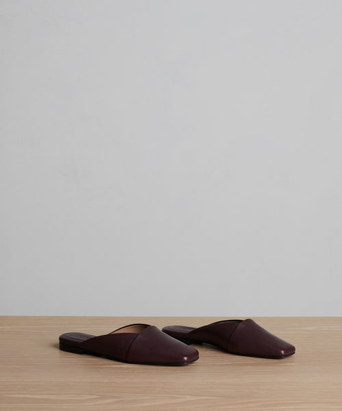 The Mia Mule: a pair of dark brown Italian leather slip-on mules with a pointed toe, displayed side by side on a light wooden surface against a plain light gray background.