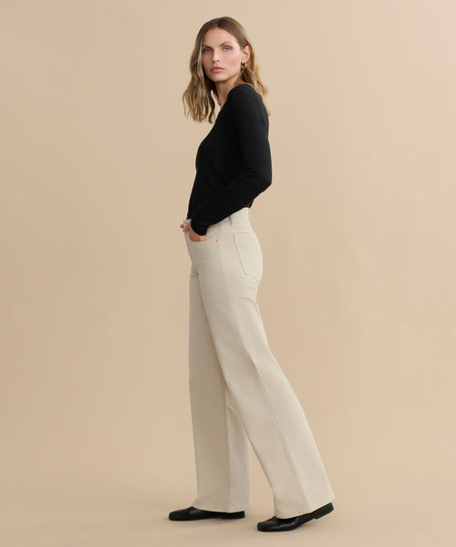 A woman stands sideways against a beige background, wearing a black long-sleeve top, Mid Rise Wide Leg pants in beige, and black shoes. She has wavy, shoulder-length hair and a neutral expression.