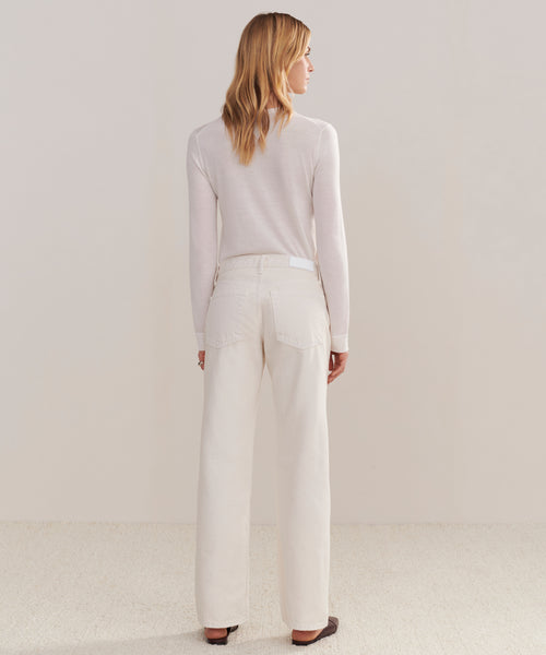 A woman faces away, wearing a long-sleeve light beige top and the Mid Rise Wide Leg jeans in winter white. She stands on a light-textured floor against a neutral background.