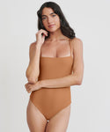 A woman with long dark hair stands against a plain white background, wearing the Malta One Piece Swimsuit in terracotta with thin straps. She has one arm crossed, the other hand touching her face, and looks at the camera.