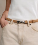 A person, visible from torso to upper legs, wears light beige jeans with hands in pockets, a white ribbed tank top, and the Mini Jules Belt—a tan calf suede belt with a distinctive rope-twist buckle.