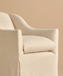A close-up of the Miramar Chair showcases its modern, cream linen slipcover, curved back, and cushioned seat, set against a beige background.