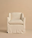 The Miramar Chair features a cream-colored linen slipcover, skirted base, plush seat cushion, and gently curved arms. Its modern, minimalist design stands out against a matching beige background.