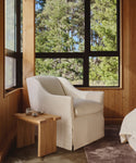 A cozy corner showcases the Miramar Chair Slipcover in white beside a wooden side table. Large windows frame lush green trees, filling the wood-paneled room with natural light.