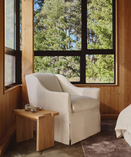 A cozy corner showcases the Miramar Chair Slipcover in white beside a wooden side table. Large windows frame lush green trees, filling the wood-paneled room with natural light.