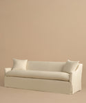 The Miramar Sofa, minimalist and cream-colored, crafted in Belgian linen with feather down fill, includes two matching pillows and is shown against a plain beige background.