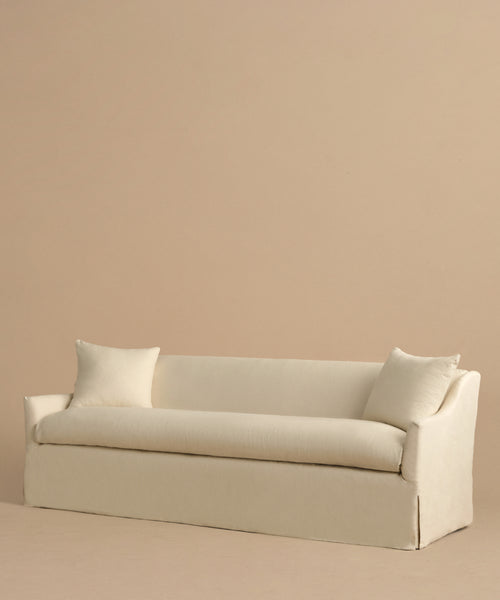 The Miramar Sofa, minimalist and cream-colored, crafted in Belgian linen with feather down fill, includes two matching pillows and is shown against a plain beige background.