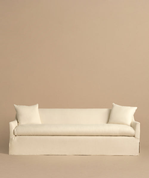 The Miramar Sofa, a minimalist cream-colored Belgian linen piece with feather down fill and two matching pillows, is set against a plain beige background for a simple, elegant look.
