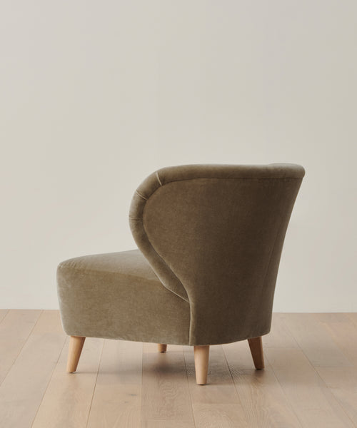 A modern, olive-green Brentwood Chair from Jenni Kayne Furniture with wooden legs sits on a light wood floor against an off-white background. The accent chair is angled to highlight its rounded back and side profile.