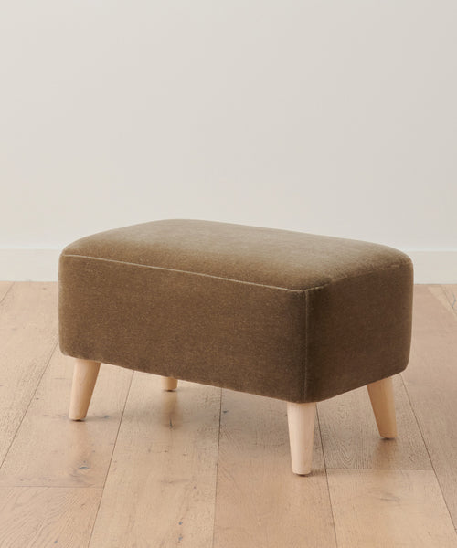 The Brentwood Ottoman is upholstered in rich brown fabric, stands on four light wood legs, and is shown on a light wood floor with a plain white wall backdrop.
