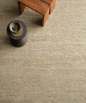 A minimalist room featuring the Mojave Jute Rug in beige, a small round black table with a bowl of three fruits, and a wooden bench nearby.