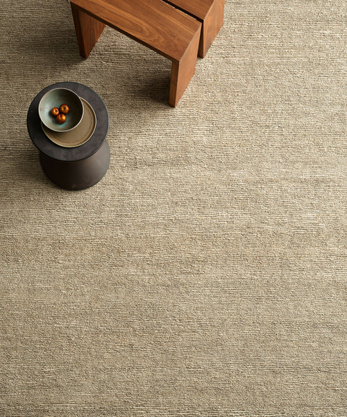 A minimalist room featuring the Mojave Jute Rug in beige, a small round black table with a bowl of three fruits, and a wooden bench nearby.