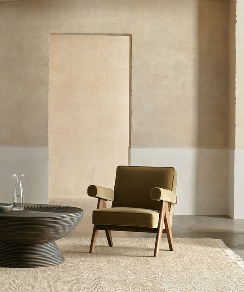 A modern living room with a mid-century olive green armchair, dark wood coffee table, clear glass carafe, neutral beige walls, and the sustainable Mojave Jute Rug handmade in India.