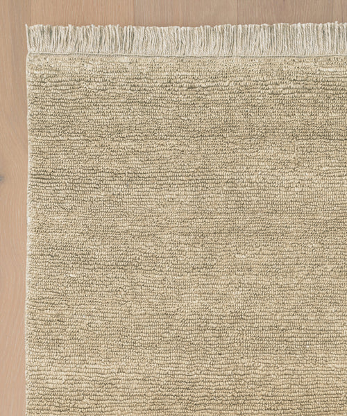 A close-up of the Mojave Jute Rug, featuring beige tones, horizontal lines, and fringed edges on a light wooden floor. Only part of the rug is visible, highlighting its handcrafted weave and detailed fringe.