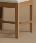 A close up of an ivory upholstered dining chair with oak wood set against a beige background.