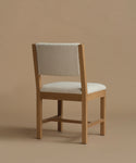 The back of an ivory upholstered dining chair with oak wood set against a beige background.