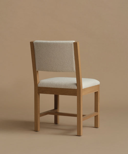 The back of an ivory upholstered dining chair with oak wood set against a beige background.