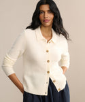 A woman with long dark hair wears the Murphy Cardigan, a fitted ivory knit, paired with a dark skirt and stands with her hands in her pockets against a neutral background.