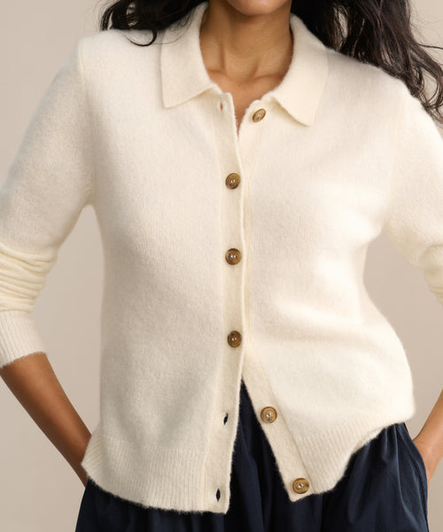 A person wearing the Murphy Cardigan, a fitted cream knit with brown buttons, over a dark skirt or pants, standing with hands in pockets.