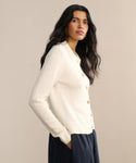 A woman with long wavy dark hair models the Murphy Cardigan, a fitted knit ivory sweater, paired with dark pants against a plain beige background, looking slightly to the side.