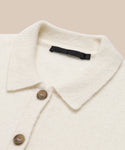 Close-up of the Murphy Cardigan—a fitted, cream wool knit with a ribbed collar, two brown buttons, and a black brand label inside the neckline—displayed on a beige background.