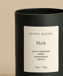 The Musk Glass Candle features a black glass vessel and is labeled jenni kayne Musk. It has spicy cardamom, amber, sandalwood, and orchid notes. Hand-poured in LA with coconut soy wax (10oz/295g), shown on a neutral beige background.
