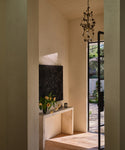 A modern interior entryway with textured beige walls and columns, featuring a black framed artwork on a light stone console table with yellow flowers, a hanging brass chandelier, and a glass door opening to a garden patio.