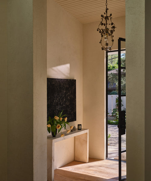 A modern interior entryway with textured beige walls and columns, featuring a black framed artwork on a light stone console table with yellow flowers, a hanging brass chandelier, and a glass door opening to a garden patio.