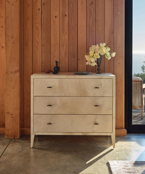 A Napa Dresser with three solid maple drawers stands against a wooden wall. A vase of white flowers and décor sit on top, while sunlight from a glass door illuminates the scene and reveals an outdoor view.