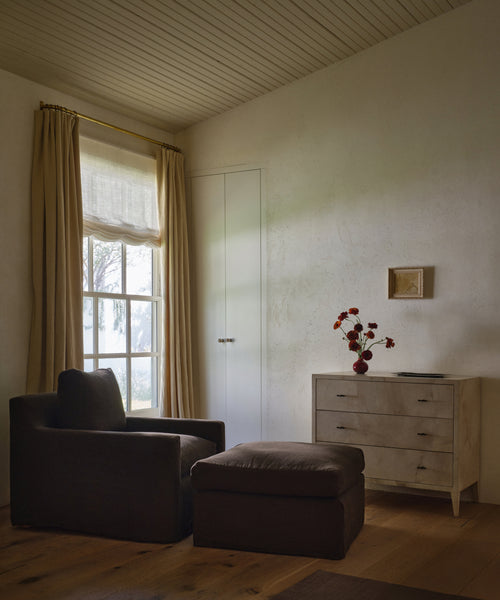 A styled interior room scene featuring a dark charcoal upholstered armchair and matching ottoman in a sunlit room with wood floors, beige walls, a window with curtains, and a light wood chest of drawers with red flowers on top.