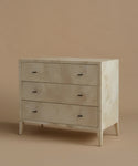 A three-drawer chest of drawers with a light beige and white marble inlay on the front and drawer fronts, supported by tapered legs, set against a solid warm beige background.