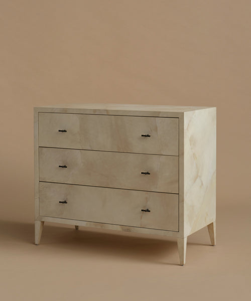 A three-drawer chest of drawers with a light beige and white marble inlay on the front and drawer fronts, supported by tapered legs, set against a solid warm beige background.