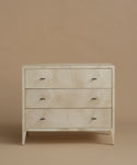 A three-drawer chest of drawers with a light beige and white marbled shell inlay and small dark metal handles, set against a solid warm beige background.