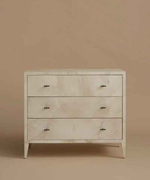 A three-drawer chest of drawers with a light beige and white marbled shell inlay and small dark metal handles, set against a solid warm beige background.