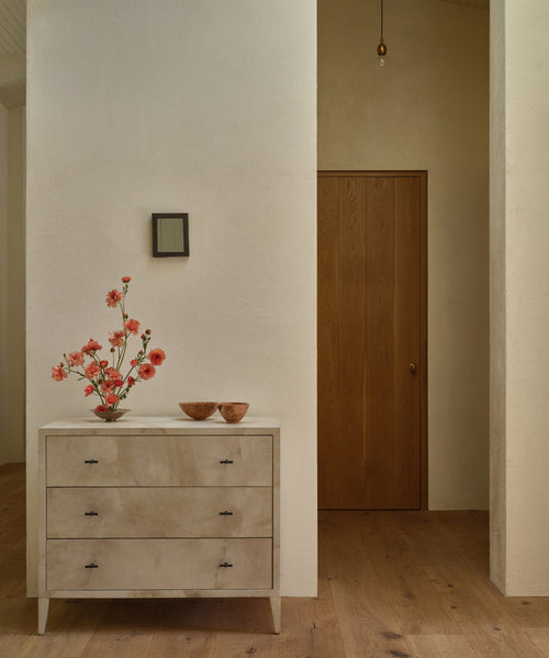 A minimalist interior showcases the Napa Dresser in light wood with solid maple drawers, topped with a vase of pink flowers and two wooden bowls. Behind are cream walls, a small square mirror, and a closed wooden door.