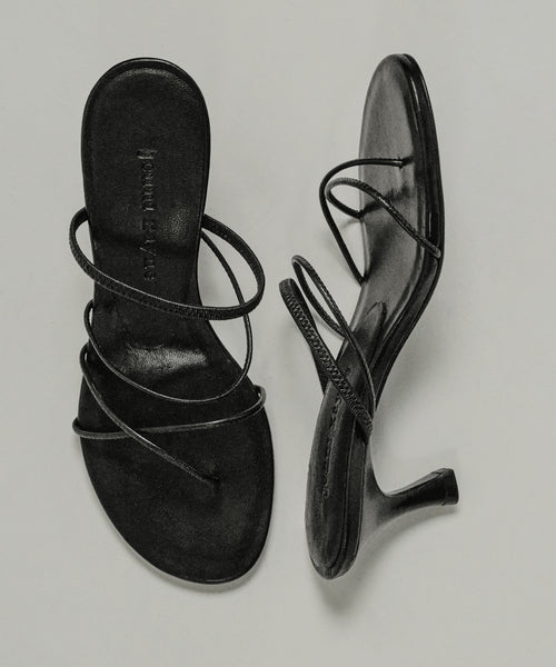 The Eve Heeled Sandal, crafted from black nappa leather with thin crisscross straps, is shown on a light background—one sandal stands upright while the other lies on its side.