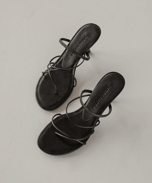 The Eve Heeled Sandal, crafted in black nappa leather with thin crisscross straps and an open toe, is displayed on a light, neutral surface.