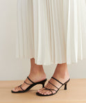 A person wearing a white pleated skirt and black Eve Heeled Sandals stands on a wooden surface.