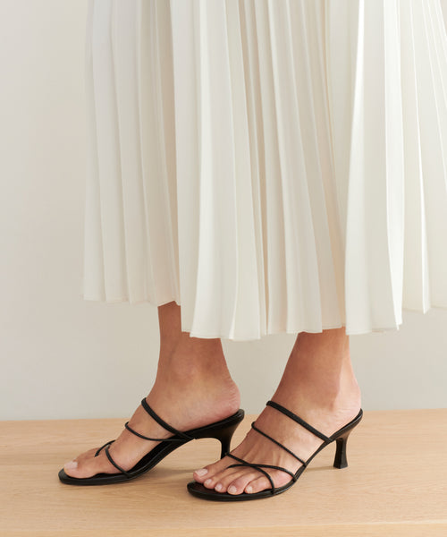 A person wearing a white pleated skirt and black Eve Heeled Sandals stands on a wooden surface.