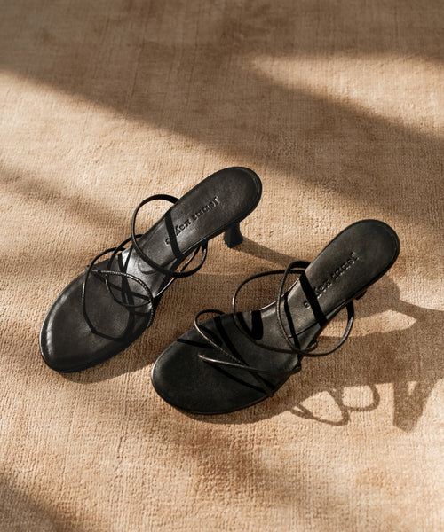 The Eve Heeled Sandal in black nappa leather, featuring elegant straps and a chic heel, is displayed on a beige textured surface with sunlight casting soft shadows over the shoes and floor.