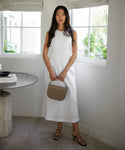 A woman stands indoors by a window, wearing a sleeveless white dress and the Eve Heeled Sandal in black, holding a taupe nappa leather handbag. The room has light wood floors, a round table, and potted plants on the windowsill.