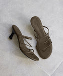 A pair of Eve Heeled Sandals in brown nappa leather with thin straps is shown on a light grey tiled floor—one sandal stands upright; the other lies flat.