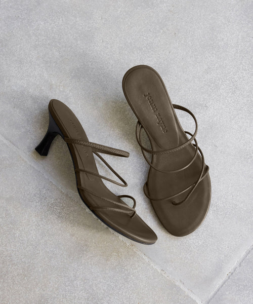 A pair of Eve Heeled Sandals in brown nappa leather with thin straps is shown on a light grey tiled floor—one sandal stands upright; the other lies flat.