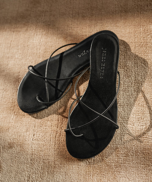 The Eve Sandal features a minimalist black design with thin crisscrossing Nappa leather straps, shown on a textured beige surface under soft natural light.
