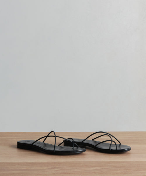 The Eve Sandal, a pair of minimalist black Nappa leather strappy sandals, sits on a wooden surface against a plain light gray background.