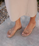 Wearing a light beige skirt, a person stands on concrete near pebbles in minimalist Eve Sandal—tan nappa leather heels featuring thin crisscross straps.